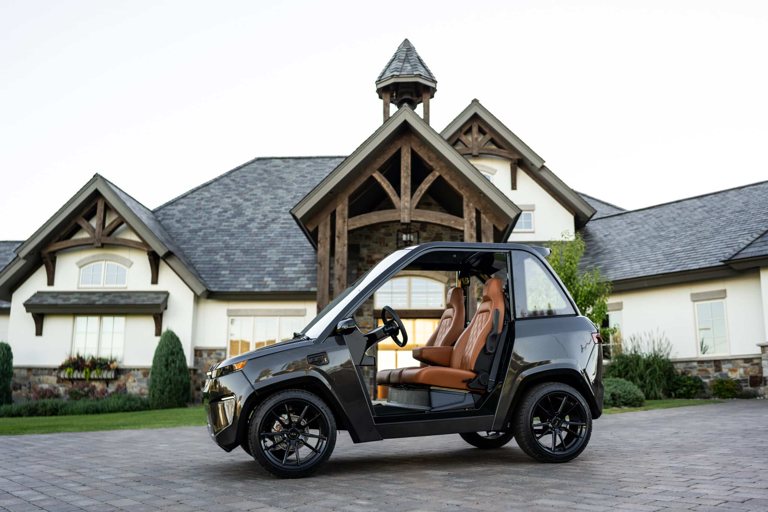 A ULV 322 in black parked in front of a large house with dark wood beams and luscious green shrubs and plants